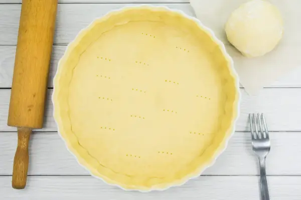 Une pâte brisée est prête dans une casserole blanche, avec une boule de pâte, une rouleau et une fourchette à côté.