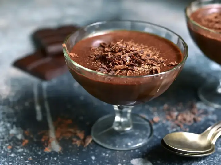 Délicieuse mousse au chocolat maison, garnie de copeaux de chocolat, dans un verre à pied. Des carrés de chocolat et une cuillère dorée complètent la scène.